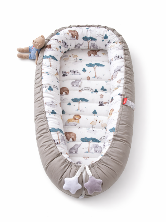 白い背景の上に、上から見たグレーのベビーネスト（ベビー用クッションベッド）が置かれている。内側は動物と木のイラスト柄で、足元は紐で絞られ星形のぬいぐるみ飾りが付いており、上部に小さなぬいぐるみが添えられている。
