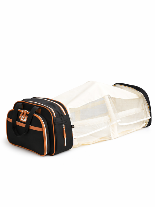 白い背景の上に、黒×キャメルのマザーズバッグと一体化したポータブルベビーベッドが横向きに置かれている。ベッド部分は白いメッシュの側面付きで、バッグには複数のファスナーポケットと持ち手が付いている。
