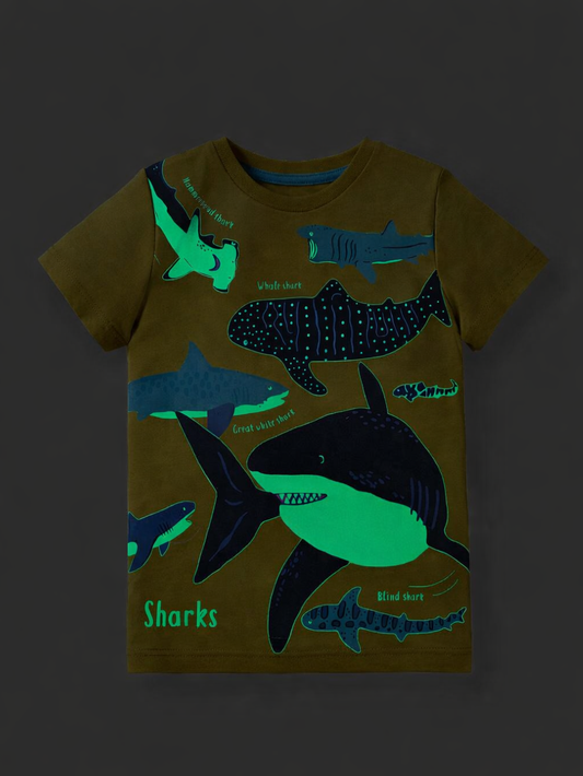 暗い背景に平置きされたキッズ用半袖Tシャツ。オリーブ色の生地に、発光するようなグリーンとブルーのサメのイラストと「Sharks」の文字がプリントされている。