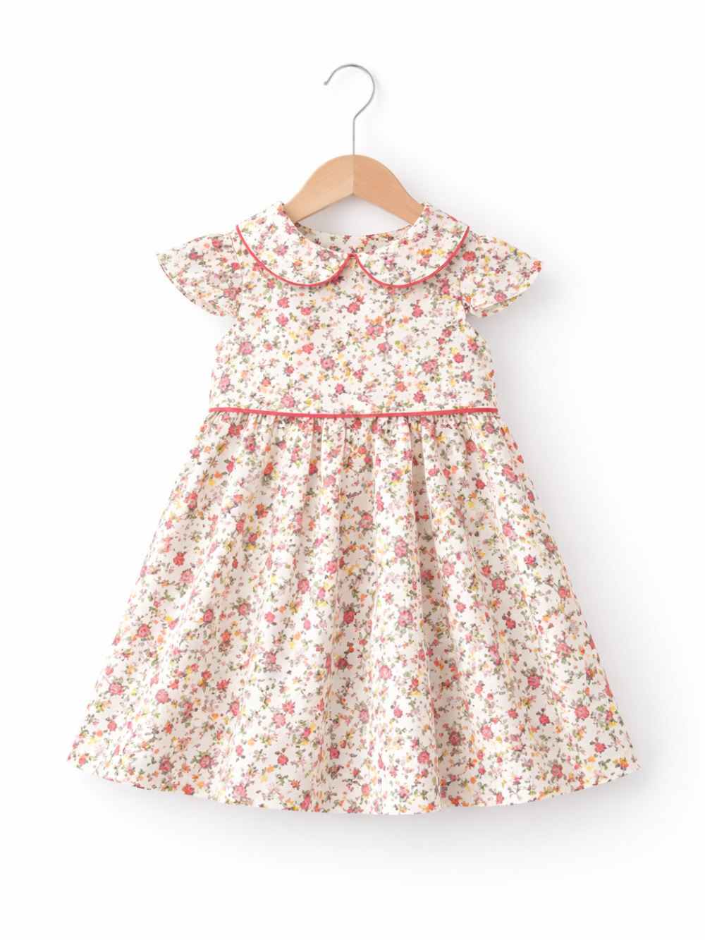 白地に小花柄の子ども用ワンピース。ピンクのパイピングが入った丸襟とフリル袖が特徴で、木製ハンガーに掛けて正面から撮影されている。