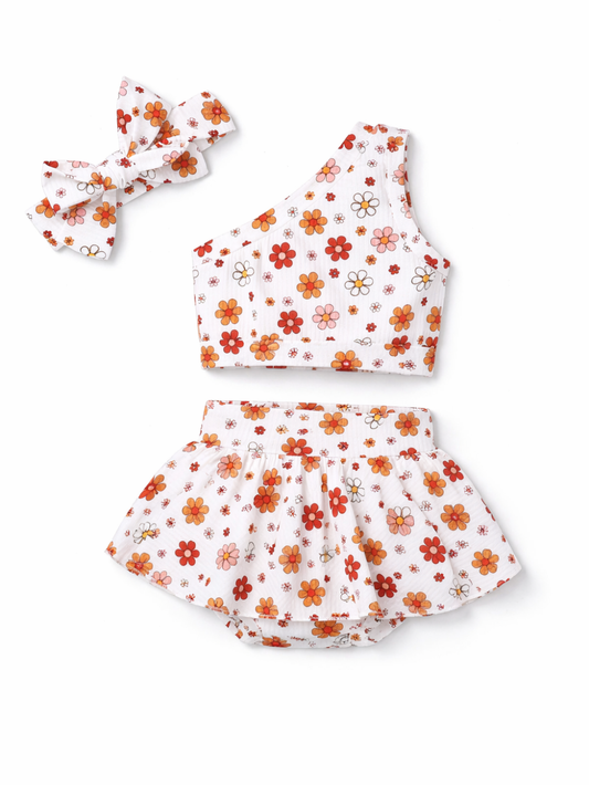 白背景に、オレンジ・赤系の花柄プリントのワンショルダートップスとフリル付きショートパンツ、同柄のリボンヘアバンドを並べた女の子用3点セット。