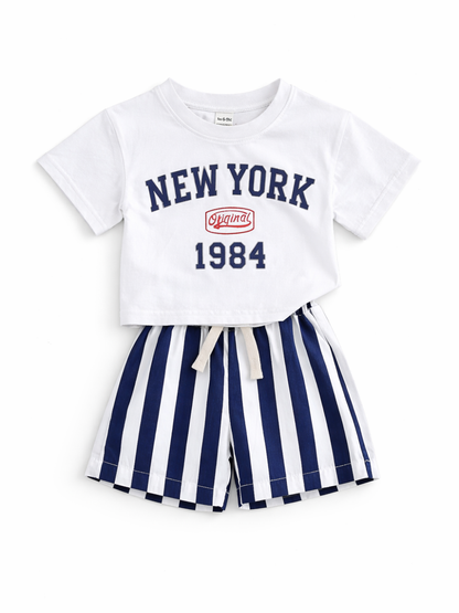 白背景に平置きされた子ども用2点セット。白い「NEW YORK 1984」ロゴ入りTシャツと、ネイビー×白の縦ストライプ柄ショートパンツ。