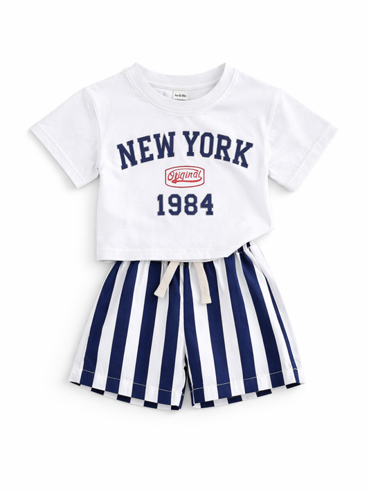 白背景に平置きされた子ども用2点セット。白い「NEW YORK 1984」ロゴ入りTシャツと、ネイビー×白の縦ストライプ柄ショートパンツ。
