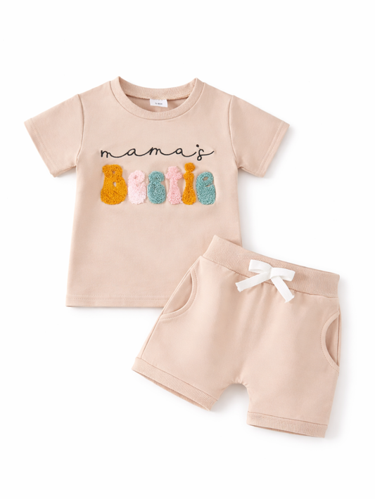 ベージュのキッズ上下セット（半袖Tシャツ＋ショートパンツ）を白背景に平置きした商品写真。Tシャツ前面に「mama’s bestie」のロゴとカラフルな立体ワッペン、パンツはウエストの白リボン付き。