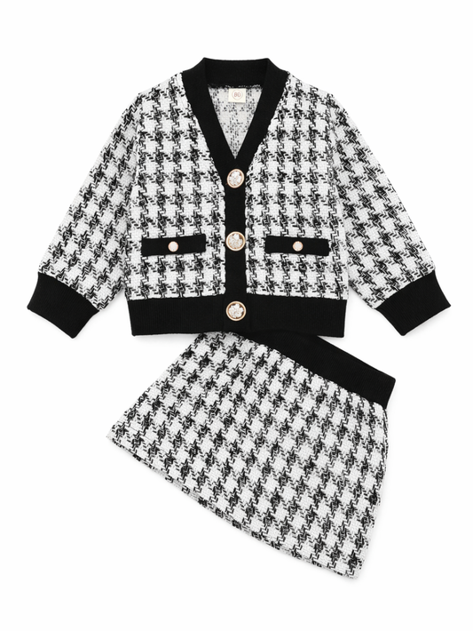 白背景の上に、黒×白の千鳥格子ツイードのキッズ用セットアップ（Vネックカーディガンとスカート）が平置きで配置されている。カーディガンは黒のリブ縁取りとゴールドボタン、胸ポケット風デザイン付き。