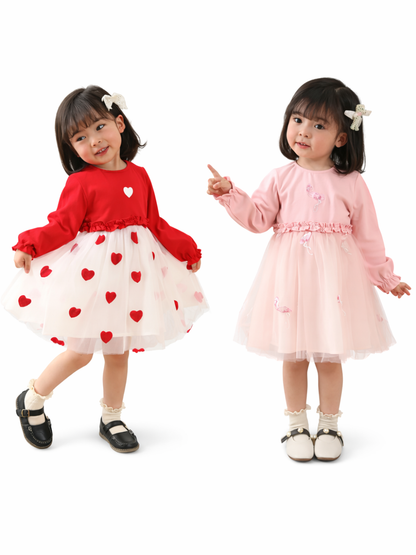 白背景のスタジオで、幼い女の子2人が並んで立ち、左は赤い長袖トップスと白いチュールスカート（赤いハート柄）、右は淡いピンクの長袖ワンピース（チュールスカート付き）を着ている。