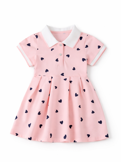 白背景のスタジオ撮影。ネイビーのハート柄が散りばめられた淡いピンクのベビー用ワンピース（白い襟・袖口、前ボタン付き）が正面から平置きで写っている。
