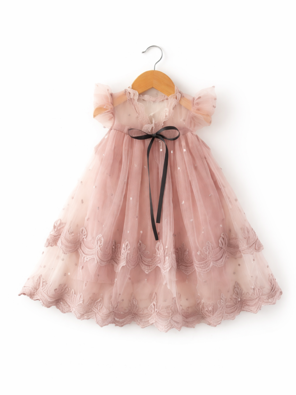 白背景に、木製ハンガーに掛かった淡いピンクのチュールワンピース。レース刺繍の裾と襟元、フリル袖、前面に黒いリボンがある。
