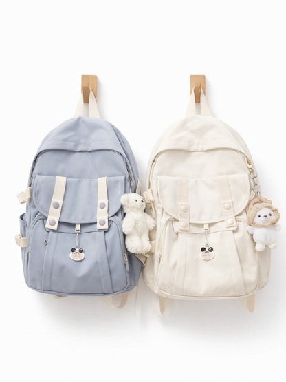 白背景に、木製フックに掛かったリュックが2つ並んでいる。左はライトブルー、右はアイボリーで、前面フラップとベルト留めのポケット、サイドポケット付き。ぬいぐるみチャームと小さなキャラクターチャームが付いている。
