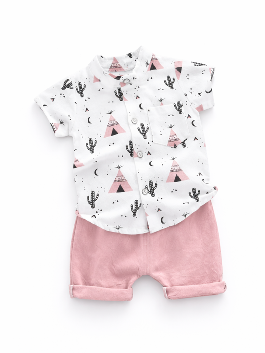 純白の背景に、子ども用の上下セットが平置きされている。サボテンやテント、三日月などの柄が入った白い半袖ボタンシャツと、淡いピンクのショートパンツの組み合わせ。

