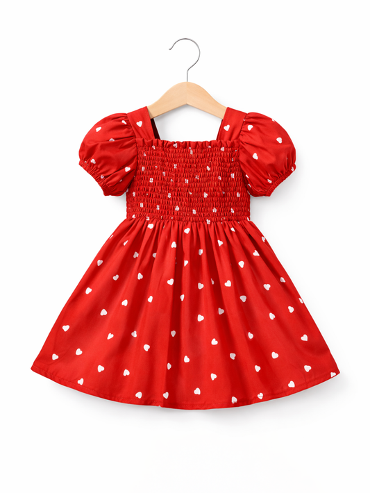 白背景に木製ハンガーで吊るした、白いハート柄の赤い子ども用ワンピース（パフスリーブ、シャーリング身頃）。
