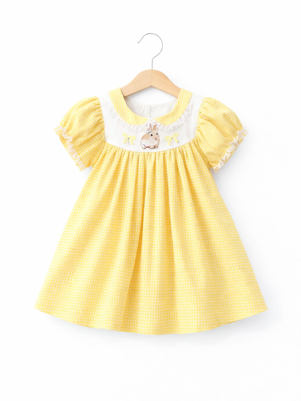 黄色のギンガムチェック柄の子ども用ワンピースが、木製ハンガーに掛けられ、純白の背景で撮影されている。パフスリーブとレース縁取り、胸元のうさぎ刺繍が特徴で、下にごく薄い自然な影がある。