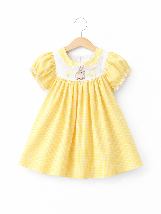 黄色のギンガムチェック柄の子ども用ワンピースが、木製ハンガーに掛けられ、純白の背景で撮影されている。パフスリーブとレース縁取り、胸元のうさぎ刺繍が特徴で、下にごく薄い自然な影がある。