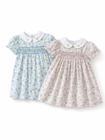 白背景に並べられたキッズワンピース2点。どちらも白い丸襟に小さな花刺繍があり、胸元はシャーリング仕様。左はブルー系の小花柄、右はピンク系の小花柄で、ふんわり広がるシルエット。
