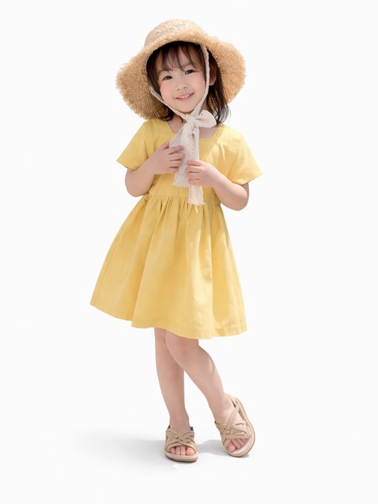 白背景で撮影された女の子の着用イメージ。麦わら帽子とレースリボンを合わせ、黄色の半袖ワンピースを着用。脚をクロスさせた上品なカタログ風ポーズで、ベージュのサンダルを履いている。
