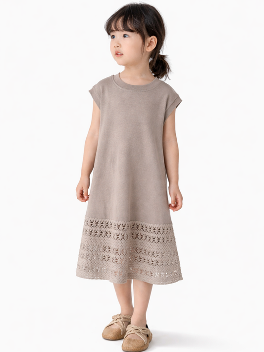 白い背景の前に立つ4〜6歳くらいの女の子。グレージュのノースリーブワンピース（裾に透かし編みレース）を着用し、ベージュのエスパドリーユ風シューズを履いている。髪は前髪ありの低めポニーテールで、正面を向いて自然に立っている。

