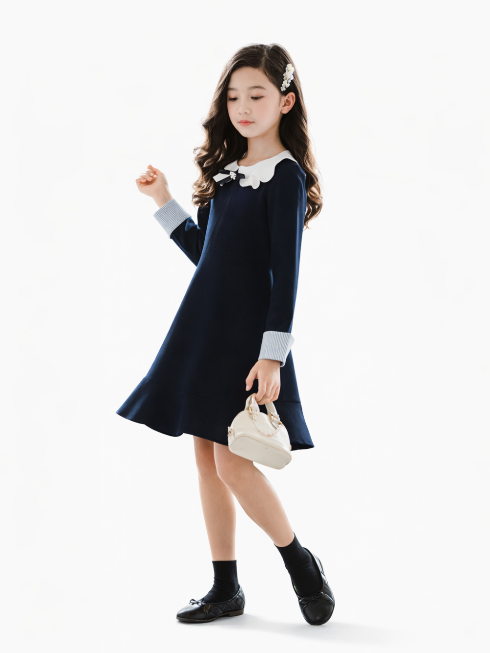 白背景のスタジオで、濃紺（ダークブルー）の長袖ワンピースを着た女の子が、クリーム色の小さなハンドバッグを持って立っている全身商品写真。
