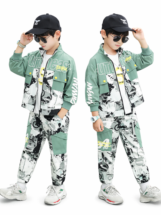 白い背景に、同じデザインのグリーン×モノクロ柄セットアップを着た男の子が2人並んで立っている。キャップとサングラスを着用し、片手で帽子のつばに触れるポーズ。ジップジャケットと総柄パンツに、イエローのアクセントと白いスニーカーを合わせている。
