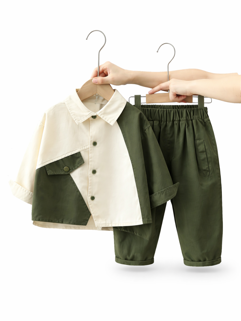白い背景に、子ども用のツートンカラーシャツとショートパンツがハンガーに掛けられて並んでいる。シャツはアイボリー×カーキの切り替えで前ボタンと胸ポケット付き、パンツはカーキのウエストゴム仕様。上部に手が写り、ハンガーを支えている。
