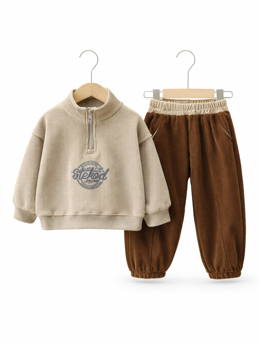 白い背景に、子ども用の上下セットがハンガーに掛けられて並んでいる。ベージュのリブ素材ハーフジップトップスと、ブラウンのコーデュロイパンツの組み合わせで、パンツはウエストゴムと裾ゴム仕様になっている。
