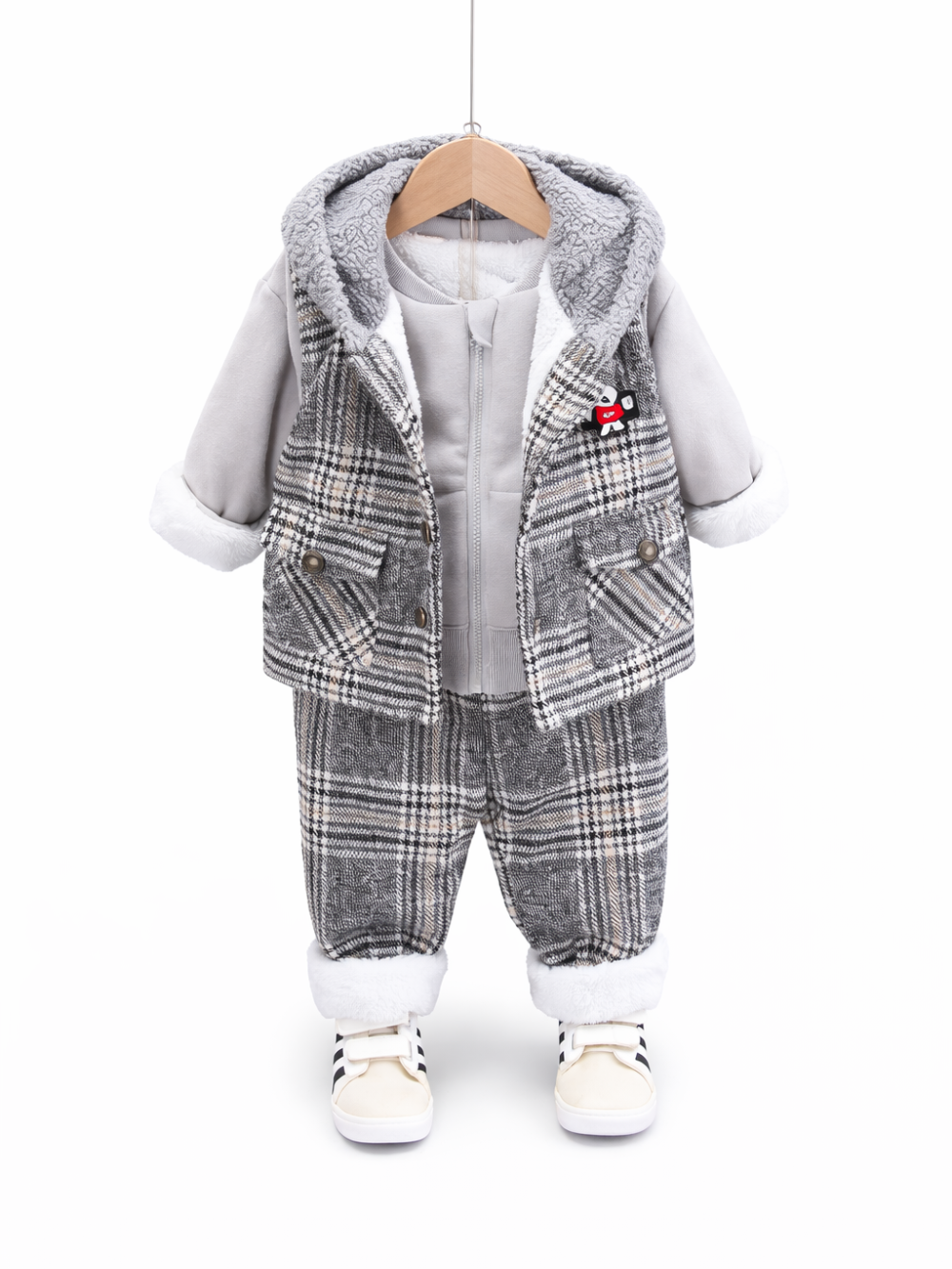 白い背景に、フード付きチェック柄ベストとグレーのジップトップス、チェック柄パンツを合わせた幼児用コーディネートがハンガーに掛けて展示されている。袖口と裾は白いボア風の縁取りで、足元にはクリーム色のスニーカーが写っている。
