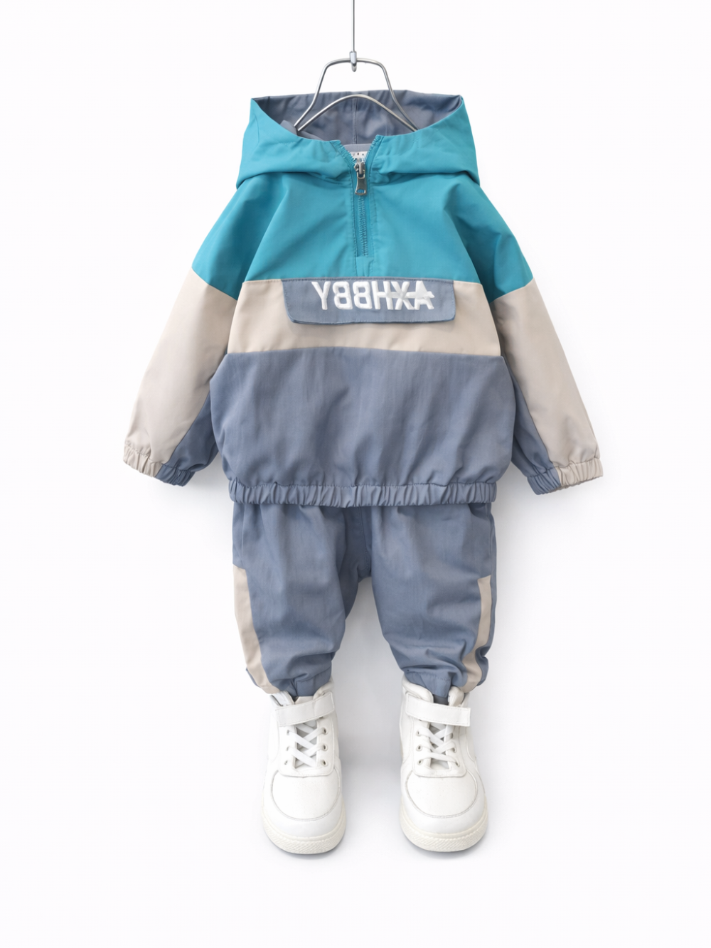 白い背景に、子ども用のフード付きジャケットとパンツの上下セットがハンガーに掛けられている。トップスはターコイズ×ベージュ×グレーの配色で、前面にロゴ入りフラップポケットがあり、パンツも同系色の切り替えデザイン。足元には白いハイカットスニーカーが写っている。

