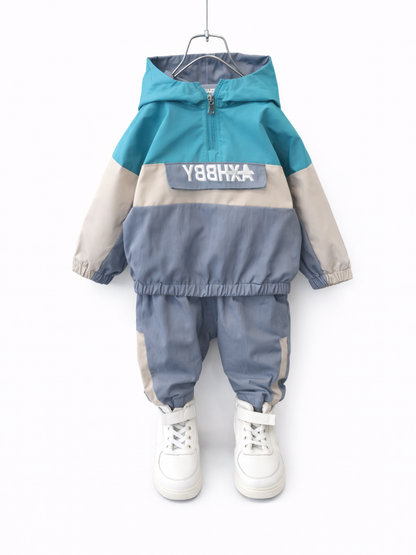 白い背景に、子ども用のフード付きジャケットとパンツの上下セットがハンガーに掛けられている。トップスはターコイズ×ベージュ×グレーの配色で、前面にロゴ入りフラップポケットがあり、パンツも同系色の切り替えデザイン。足元には白いハイカットスニーカーが写っている。

