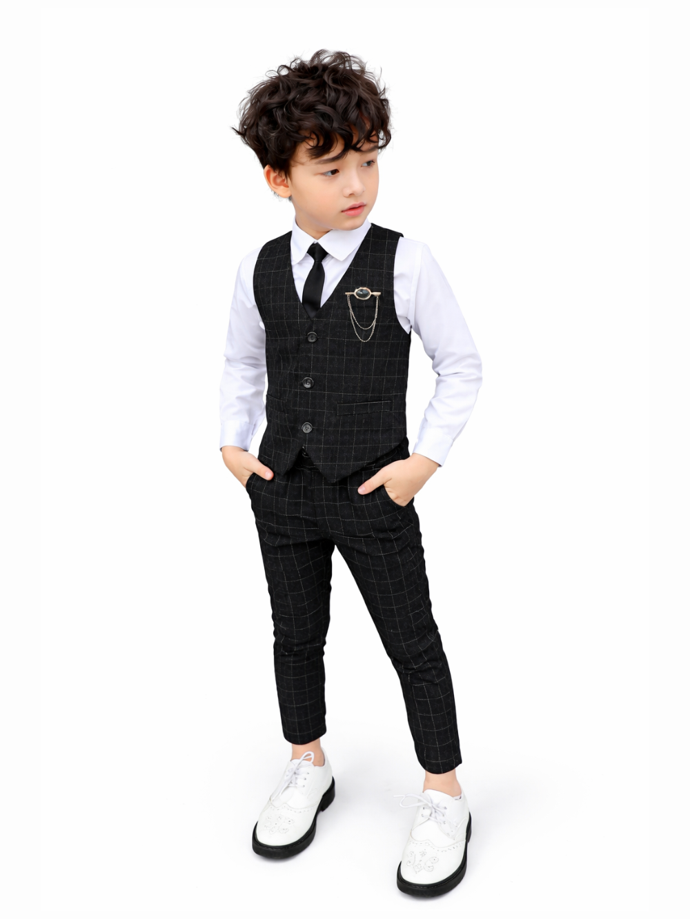 白い背景に、チェック柄のベストとパンツを合わせたフォーマルなスーツスタイルの男の子が立っている。白いシャツにブラックのネクタイを締め、ベスト胸元にチェーン付きブローチが付いている。白いシューズを履き、両手をポケットに入れて横を見ている。
