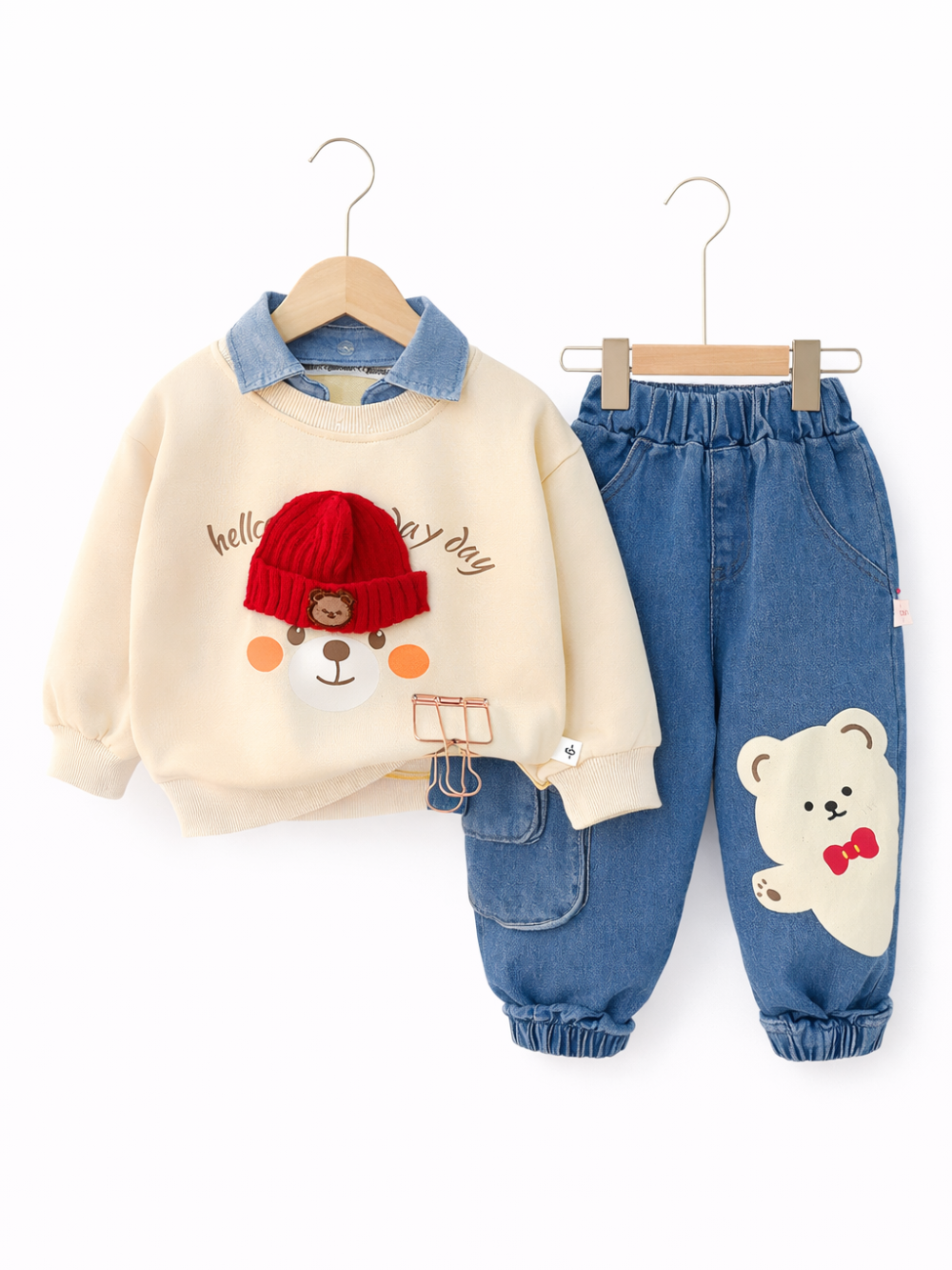 白い背景に、子ども用のトレーナーとデニムパンツの上下セットがハンガーに掛けられて並んでいる。トレーナーはアイボリーでデニム風の襟付き、前面に赤いニット帽をかぶったクマのイラストがある。パンツはブルーデニムで、脚にクマのアップリケが付いている。
