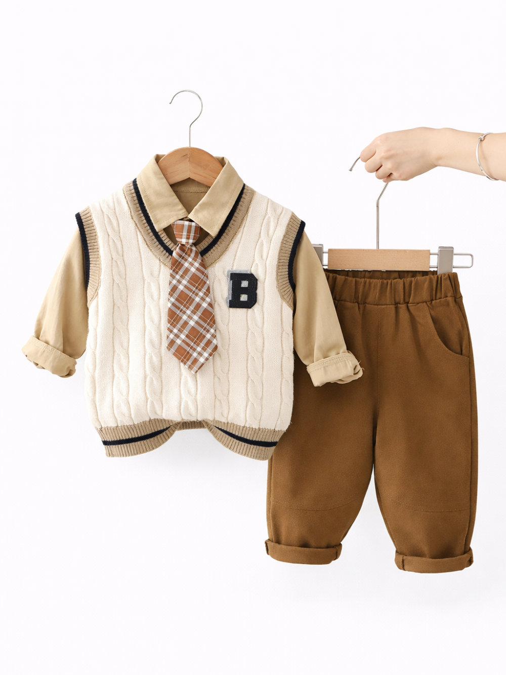 ベージュのシャツにケーブル編みのクリーム色ニットベストとチェック柄ネクタイを合わせた子ども服の上衣と、ブラウンのパンツがハンガーに掛けられ、白背景で撮影されている。
