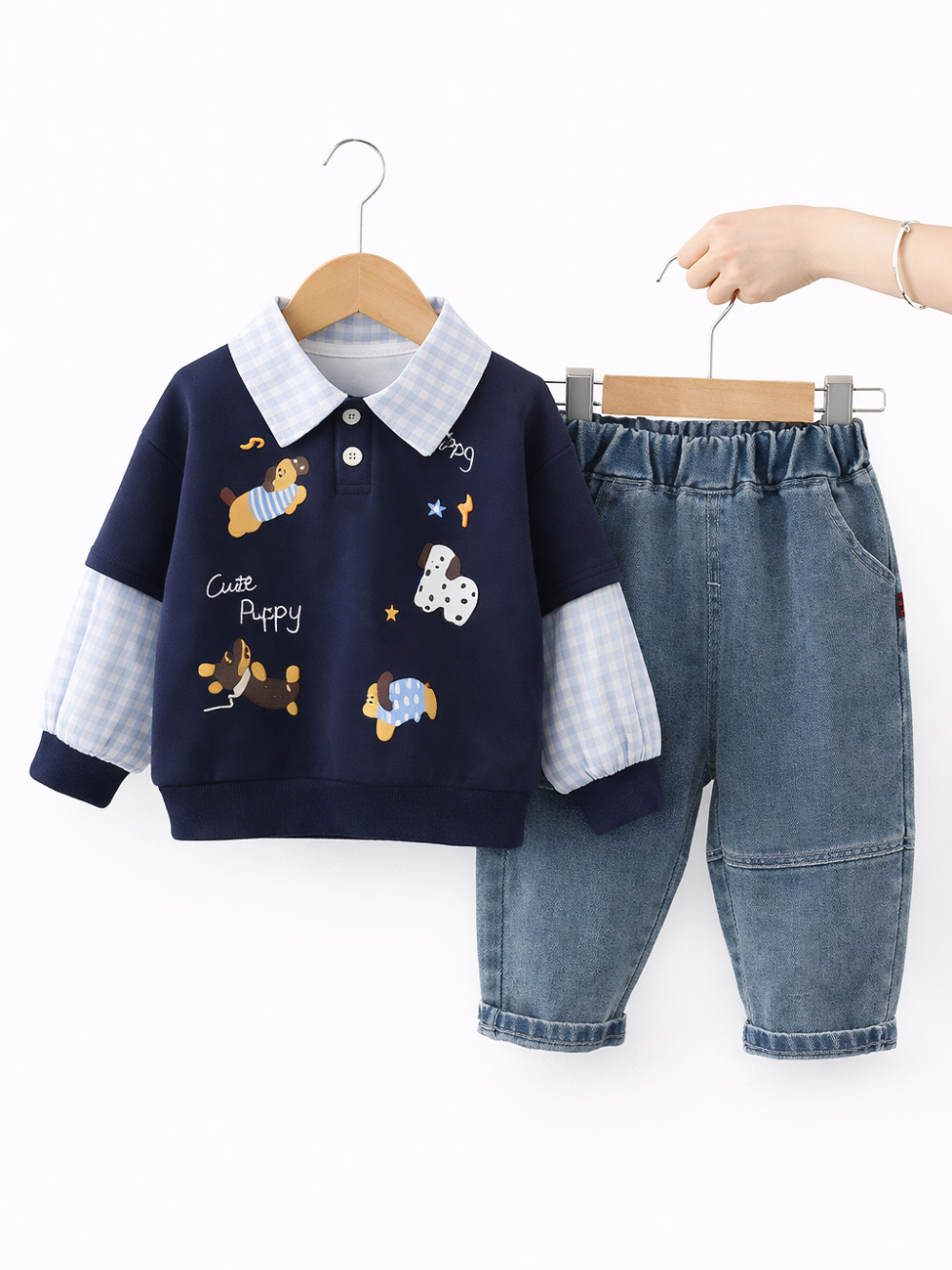 ネイビーのキッズ用長袖トップス（ギンガムチェックの襟と袖、犬のイラスト入り）と、ブルーデニムのパンツが木製ハンガーに掛けられ、白い背景の前で並んでいる。右側にはパンツのハンガーを持つ手が写っている。
