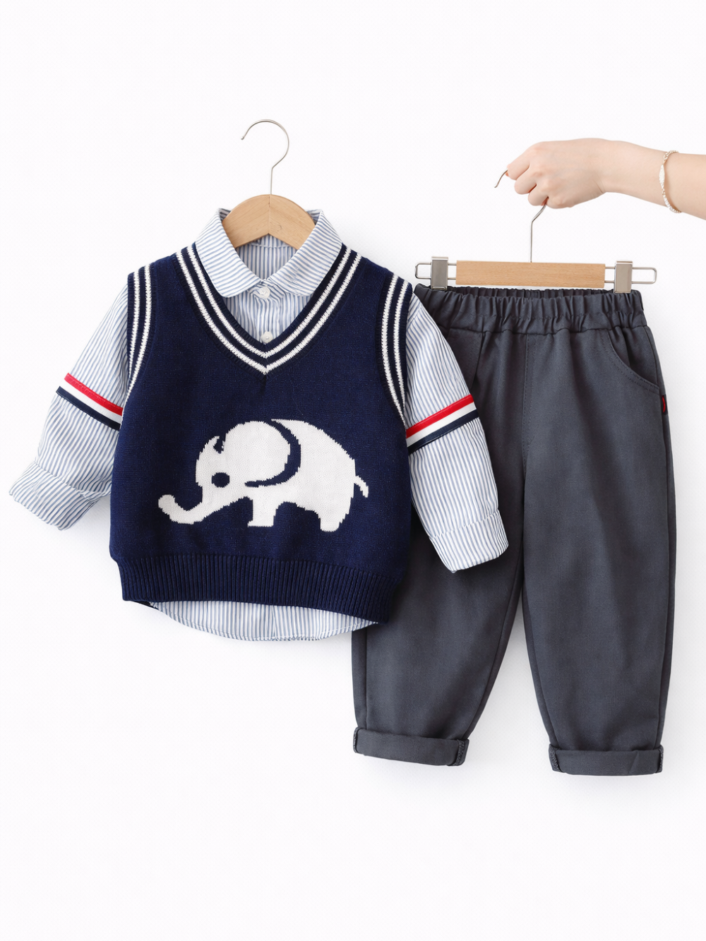 白い背景の前で、子ども服のコーディネートがハンガーに掛けられている。ネイビーのVネックニットベストには白いゾウの柄が入り、下に青白ストライプのシャツを重ねている。右側にはチャコールグレーのパンツが並び、手がハンガーを支えている。
