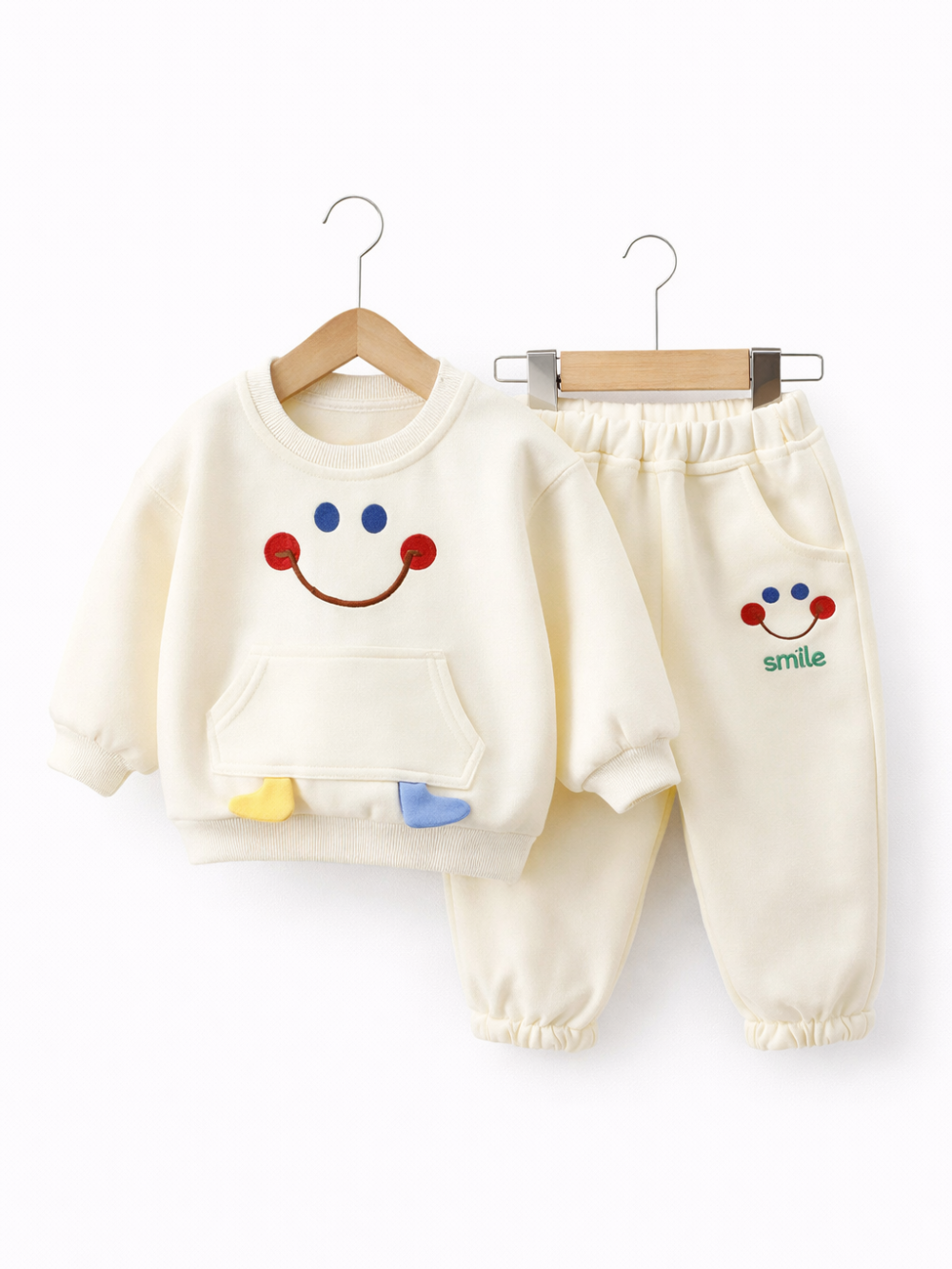 クリーム色のキッズ用上下セット（スウェット＋ジョガーパンツ）がハンガーに掛けられ、白背景のスタジオで並んで写っている。トップスは笑顔のモチーフ、パンツには小さな笑顔と「smile」の文字がある。

