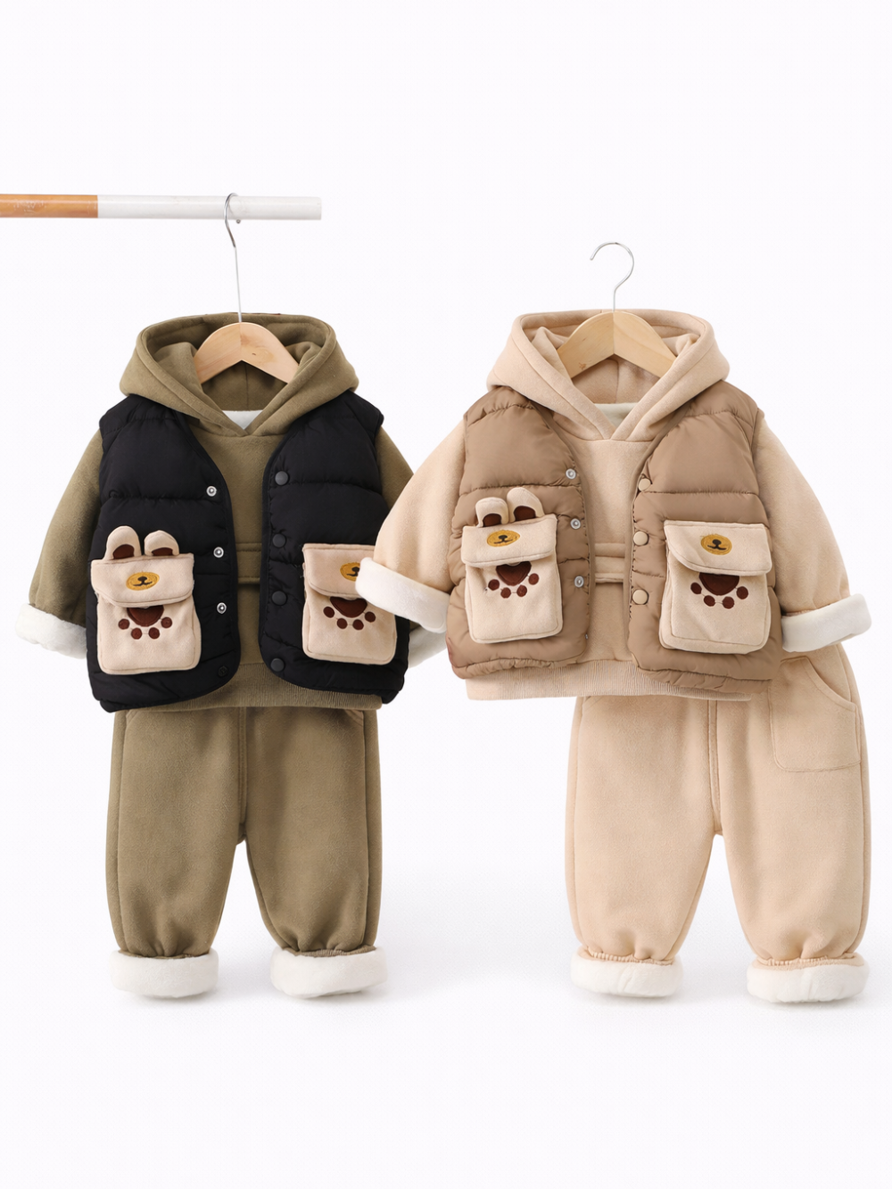 白背景のスタジオ風に、子ども用の冬服セット2点がハンガーに掛けて並んでいる。左はカーキのフーディーに黒の中綿ベスト、右はベージュのフーディーにベージュの中綿ベストで、どちらもくまモチーフのポケット付き。下は裏起毛風のスウェットパンツで、裾は白いボア仕様。