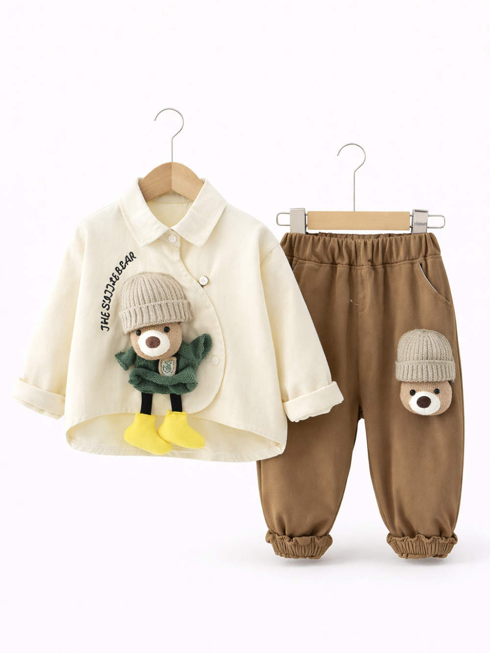 白い背景の前で、木製ハンガーに掛けられた子ども用の上下セット。左にクリーム色のシャツ（胸元に「THE SUDDER BEAR」の刺繍と立体のくまモチーフ）、右にブラウンのパンツ（太ももにくまの顔モチーフ）。