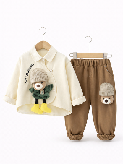 白い背景の前で、木製ハンガーに掛けられた子ども用の上下セット。左にクリーム色のシャツ（胸元に「THE SUDDER BEAR」の刺繍と立体のくまモチーフ）、右にブラウンのパンツ（太ももにくまの顔モチーフ）。