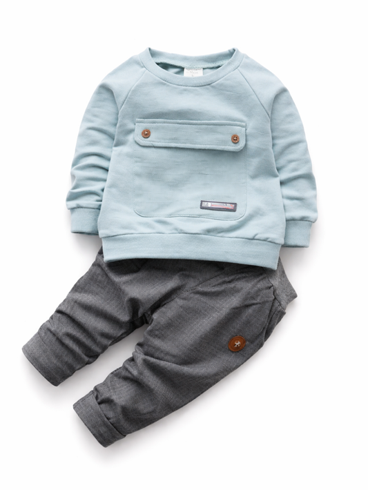 白背景に置かれた、ライトブルーのスウェットトップスとチャコールグレーのパンツのベビー用上下セット。胸に大きなポケットとボタン付きで、パンツはロールアップ裾。
