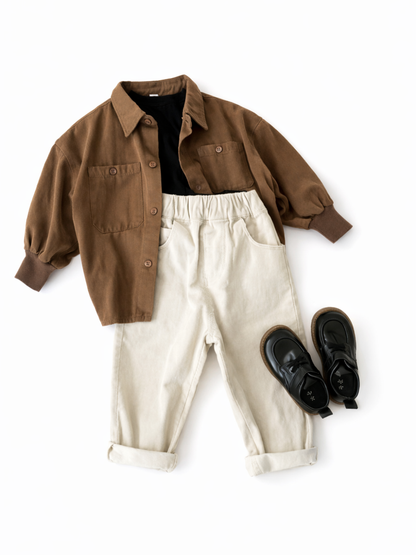 ベージュのラグの上に並べられた子ども服のフラットレイ。ブラウンのシャツジャケットとアイボリーのパンツ、黒い靴とロゴ入りの紙袋が置かれている。
