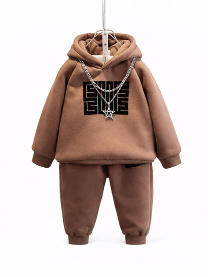 白背景にハンガーで吊るされたブラウンのキッズ用フーディーとジョガーパンツのセット。胸に黒いロゴがあり、シルバーチェーンの星形ペンダントが掛かっている。
