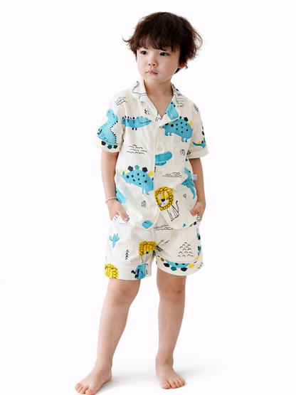 白背景のスタジオで、恐竜とライオン柄のクリーム色パジャマ上下を着た男の子がポケットに手を入れて立っている全身写真（裸足）。
