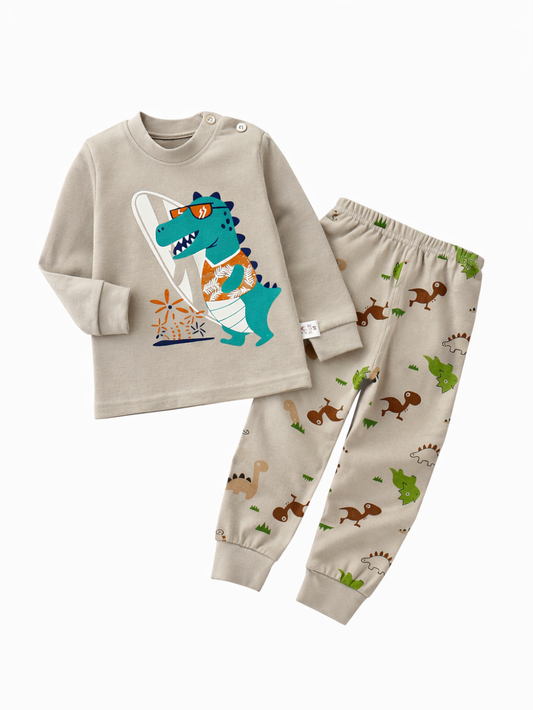 ベージュ系の長袖トップスとロングパンツの子ども用上下セット。トップスはサーフボードを持った恐竜プリントで、肩にボタン付き。パンツは恐竜柄の総柄で、白背景のスタジオ風に並べて撮影されている。
