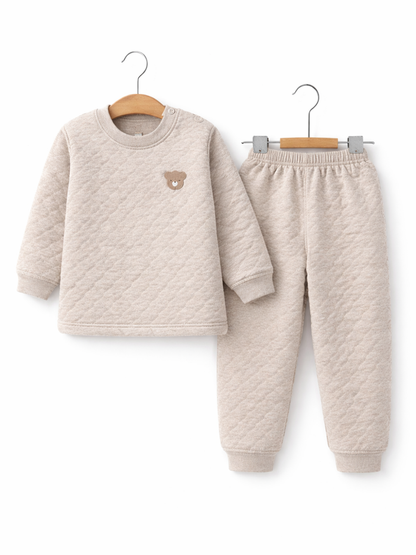 ライトベージュのキッズ用キルト素材ルームウェア上下セット。ダイヤ柄の長袖トップス（胸に小さなクマ刺繍）と、同素材のロングパンツがそれぞれハンガーに掛けられ、白背景のスタジオで撮影されている。
