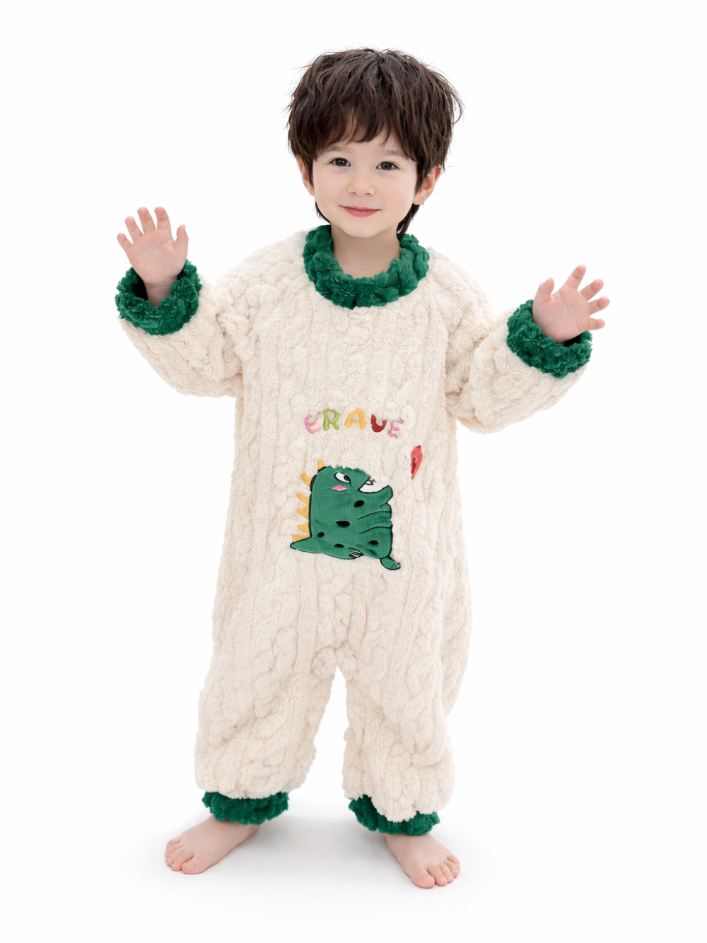 白背景のスタジオ風に、クリーム色のふわふわつなぎパジャマを着た幼児が立って微笑んでいる。襟・袖口・裾はグリーンのボアで、胸に「BRAVE」の刺繍と恐竜アップリケが入っている。
