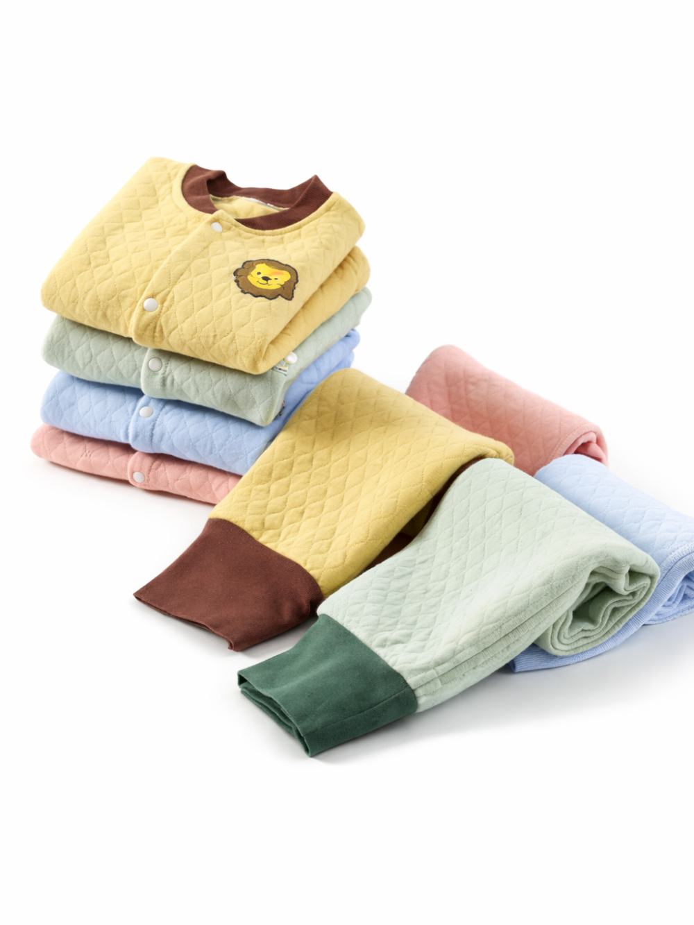 白背景のスタジオ風に、キルト素材のベビー服セットがカラー違いで重ねて並べられている。ライオンワッペン付きのイエロー上着を含むパステルカラーのトップスが積まれ、前には同素材のパンツ（イエロー×ブラウン、ミント×グリーン、ブルー、ピンク）が折りたたんで配置されている。
