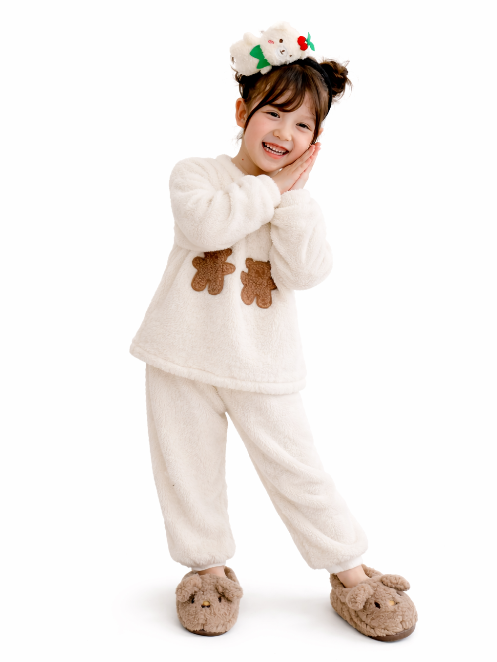 白いふわふわ素材のルームウェアを着た女の子が、白背景のスタジオで笑顔で手を頬に添えて立っている。くまのアップリケ付きトップスとパンツ、くまのスリッパ、髪はお団子でヘアアクセサリーを着用。
