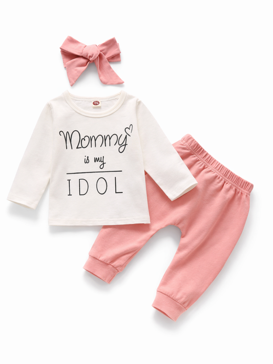 白背景に、ベビー用の3点セット（白い長袖トップス、ピンクのパンツ、ピンクのリボンヘアバンド）が並んでいる。トップスには「Mommy is my IDOL」とハートのプリントが入っている。
