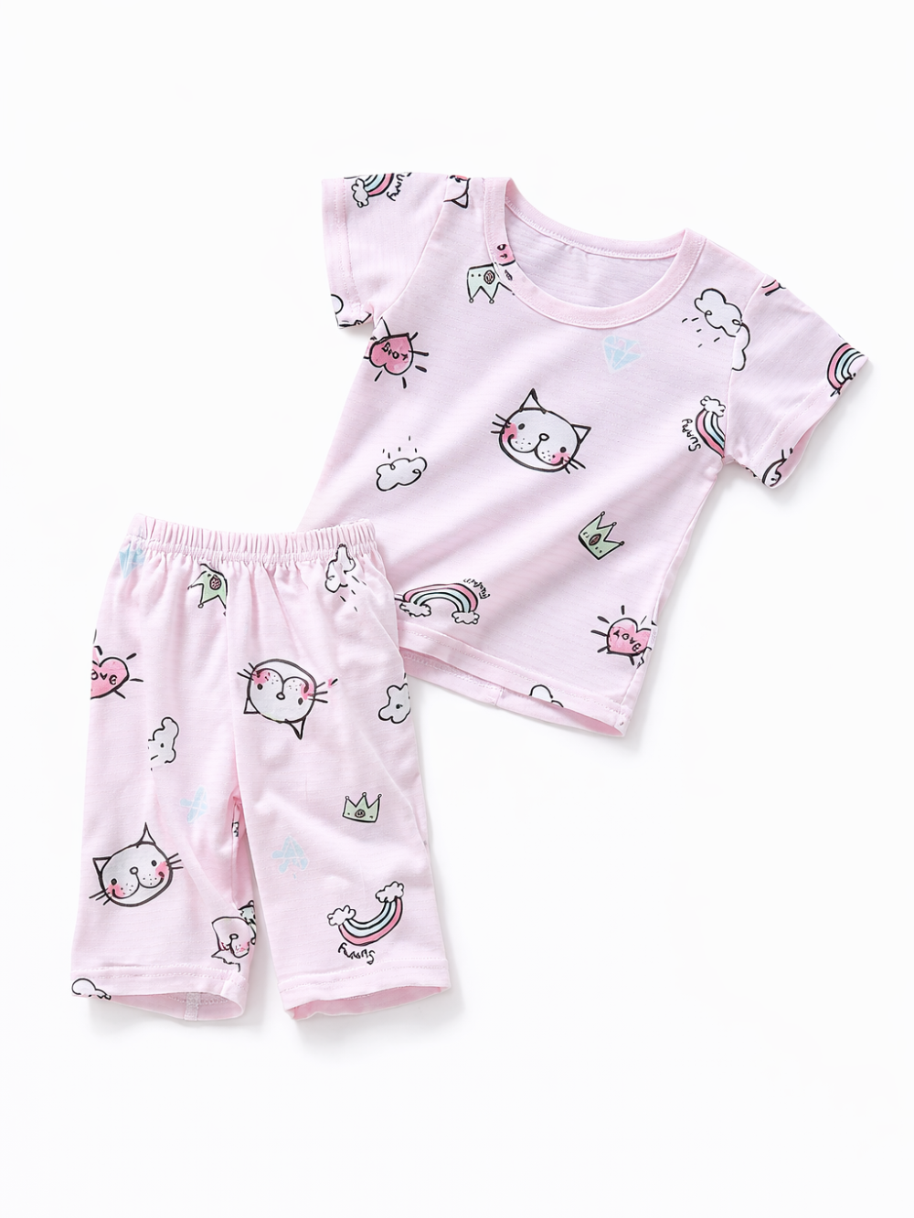 淡いピンクの半袖Tシャツと七分丈ショーツのキッズパジャマセット。猫の顔、虹、雲、王冠などのポップな総柄プリントで、白背景に斜め配置されている。
