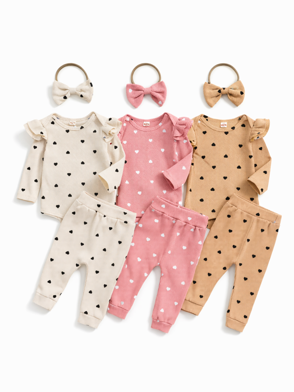 リブ素材のベビー用上下セット3色（アイボリー・ピンク・ベージュ）を白背景に平置きした商品写真。ハート柄の長袖ロンパースとパンツ、リボンヘアバンドが各色で揃っている。
