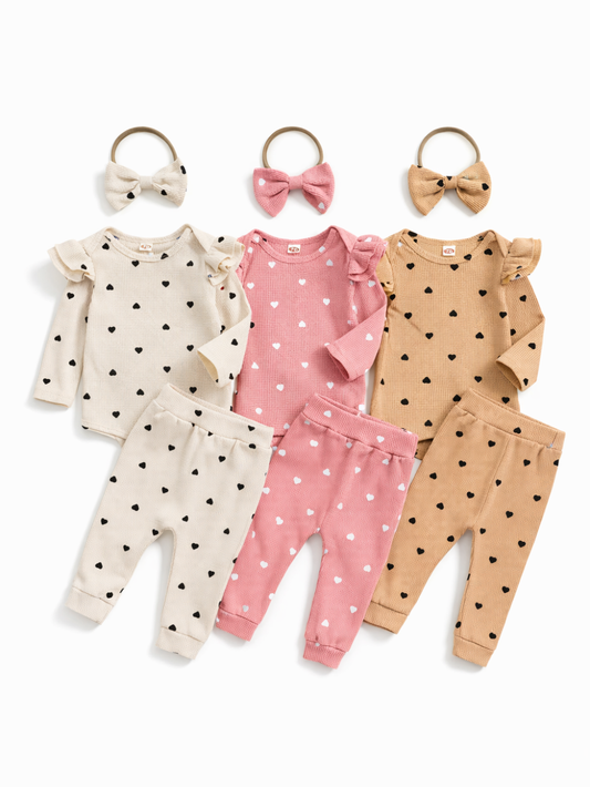 リブ素材のベビー用上下セット3色（アイボリー・ピンク・ベージュ）を白背景に平置きした商品写真。ハート柄の長袖ロンパースとパンツ、リボンヘアバンドが各色で揃っている。
