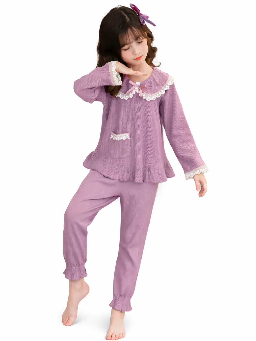 白背景のスタジオで、レース付きの襟と袖口があるラベンダー色の長袖トップスとパンツのルームウェアを着た女の子が、髪に紫のリボンを付けて裸足でポーズをとっている。
